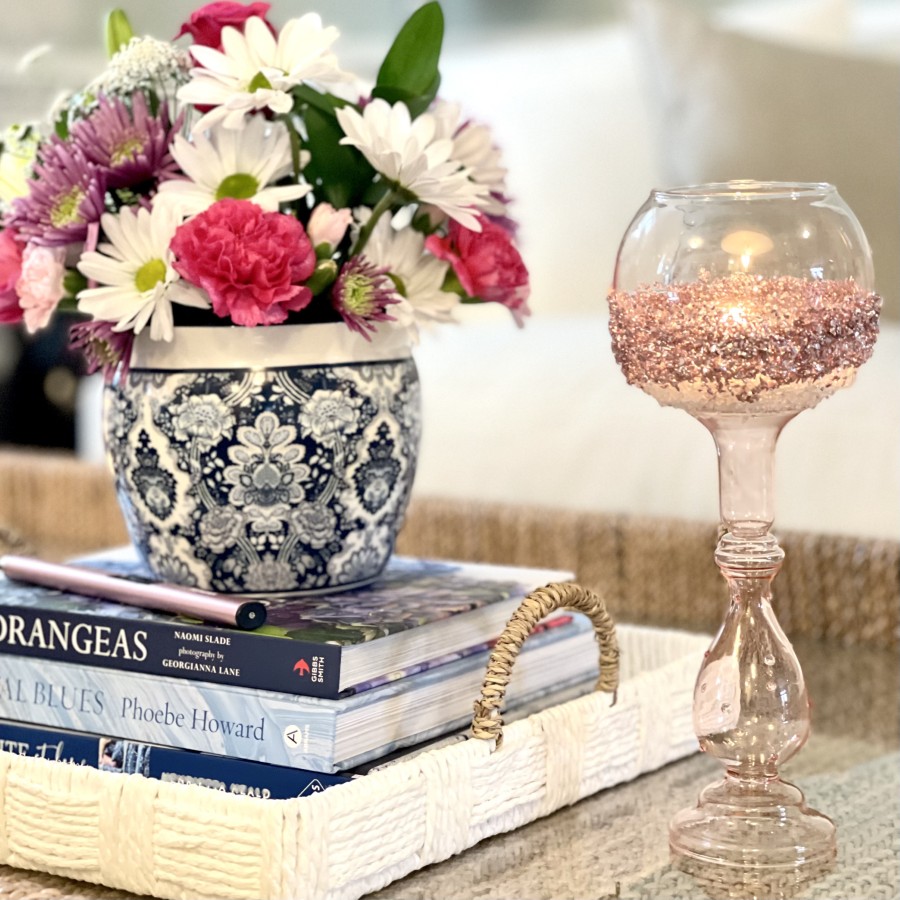 A woven tray holds a floral arrangement in a blue and white vase, alongside a glass candle holder with a textured base. Several decorative books are stacked beneath the vase on a light-colored table surface.