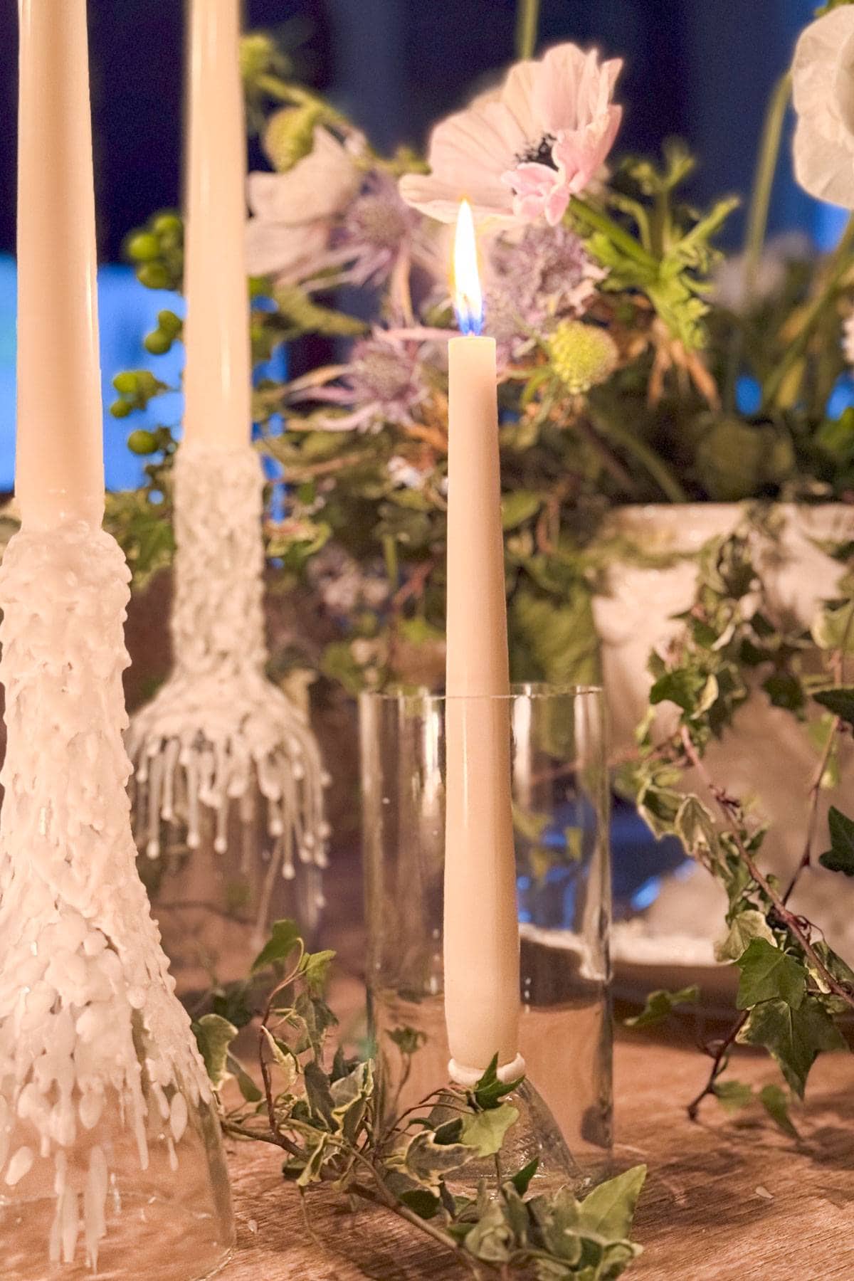 A lit candle in a glass holder is surrounded by greenery and flowers. Two other candles, with wax dripped down their sides, stand nearby. Soft lighting highlights the arrangement, creating an elegant and warm atmosphere.