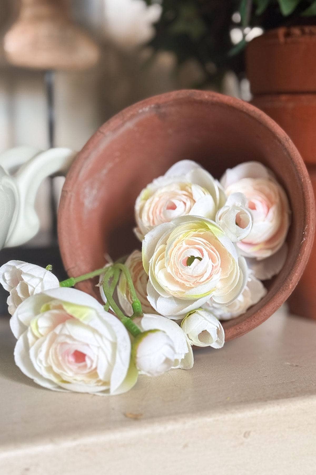 A terracotta pot lays on its side, with several cream and pale pink artificial flowers spilling out. There is a green stem attached to the flowers, and a second pot partially visible in the background.