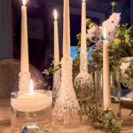 Five lit taper candles with wax drippings are arranged on a table among green foliage and a floral arrangement. A single floating candle is in a clear glass container in the foreground. The setting is dimly lit, creating a warm ambiance.