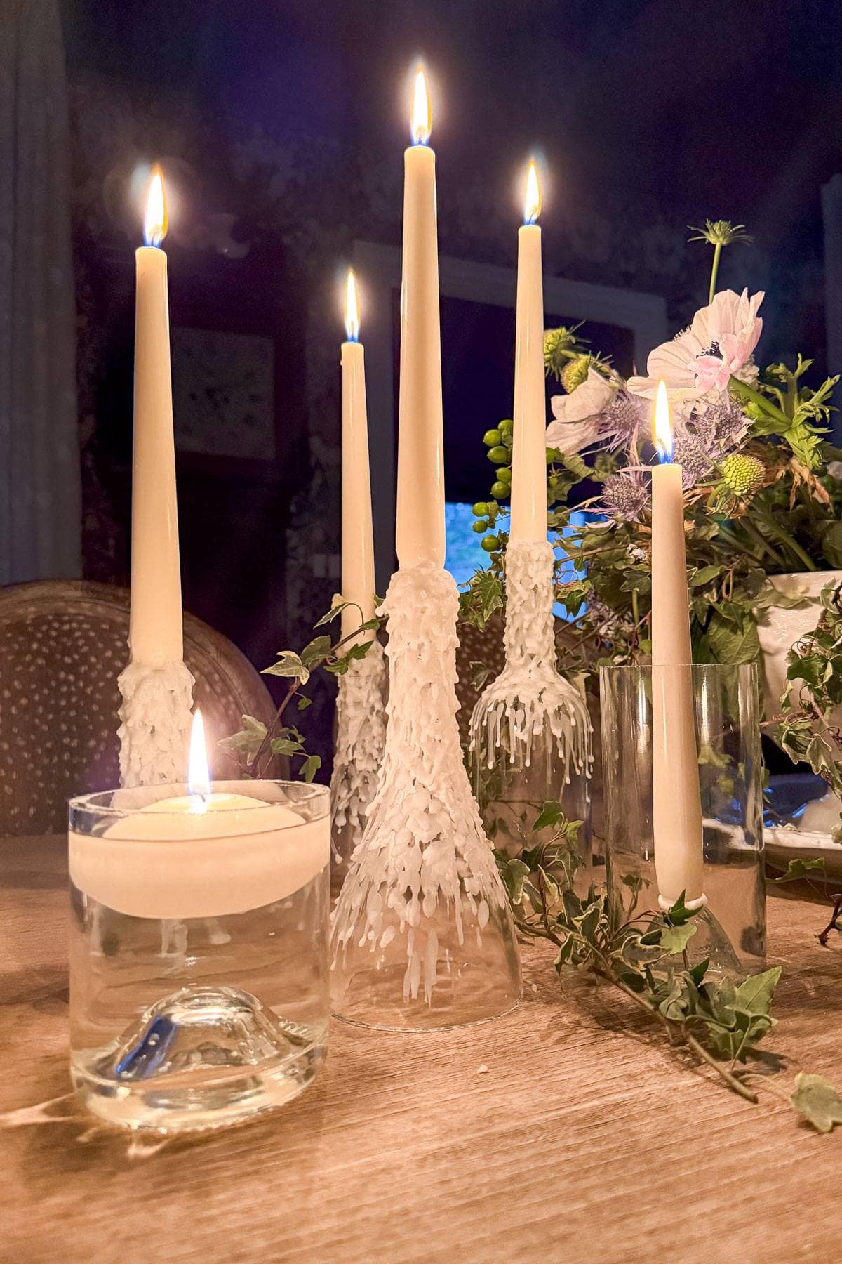 Five lit taper candles with wax drippings are arranged on a table among green foliage and a floral arrangement. A single floating candle is in a clear glass container in the foreground. The setting is dimly lit, creating a warm ambiance.