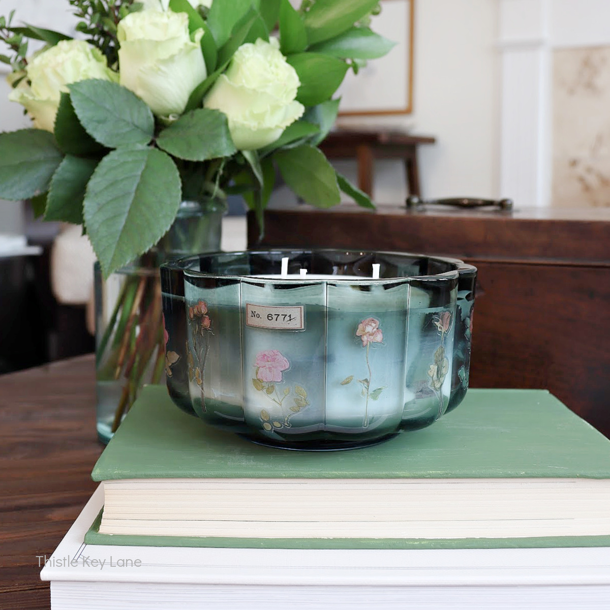 A decorative candle with floral designs sits on a stack of green books. Fresh white roses in a vase are in the background, adding a touch of elegance to the scene. The setting appears cozy and inviting.