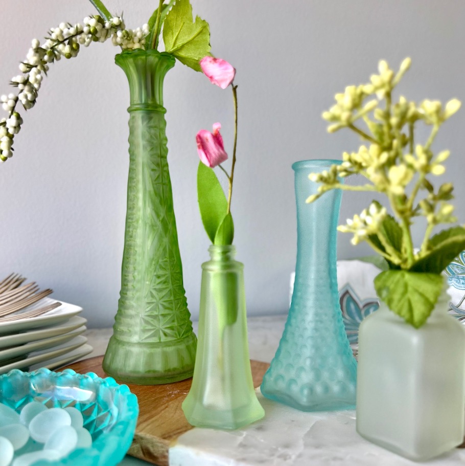 A collection of green and blue glass vases in varied shapes and sizes, each holding small, delicate flowers. The scene includes a stack of plates and a bowl with textured glass pebbles on a marble surface.