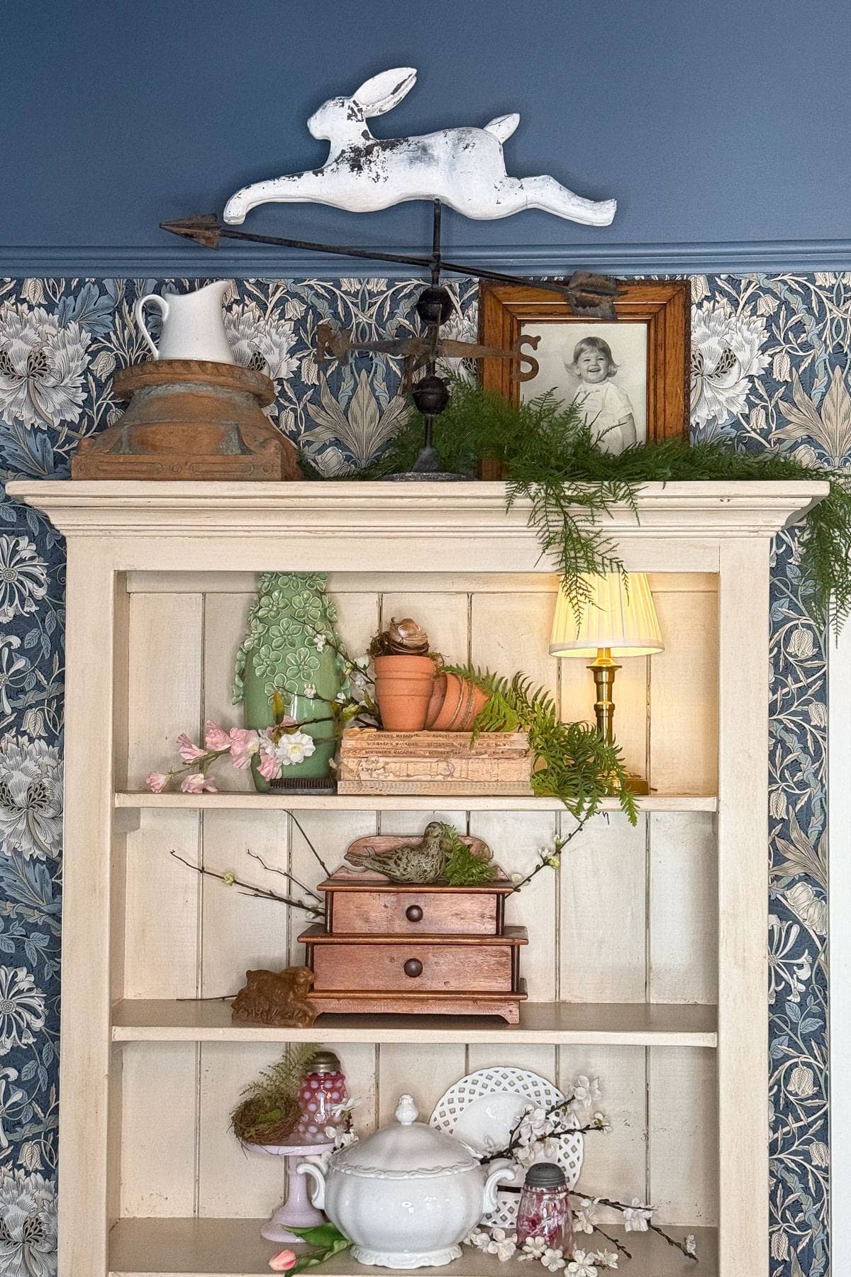 A white shelf adorned with decorative items: pottery jug, vintage rabbit weathervane, framed photo, terracotta pots, greenery, pink blossoms, small drawers, white teapot, pink cups, and a plate. Blue floral wallpaper background with a small lamp.