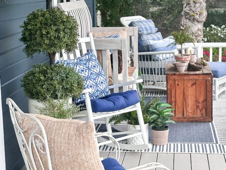 A cozy porch featuring white rocking chairs with blue and patterned cushions, a wooden coffee table, potted plants, and a wicker chair. The porch floor is covered with a striped rug, perfectly framing the greenery in the background—a perfect pour for relaxation.