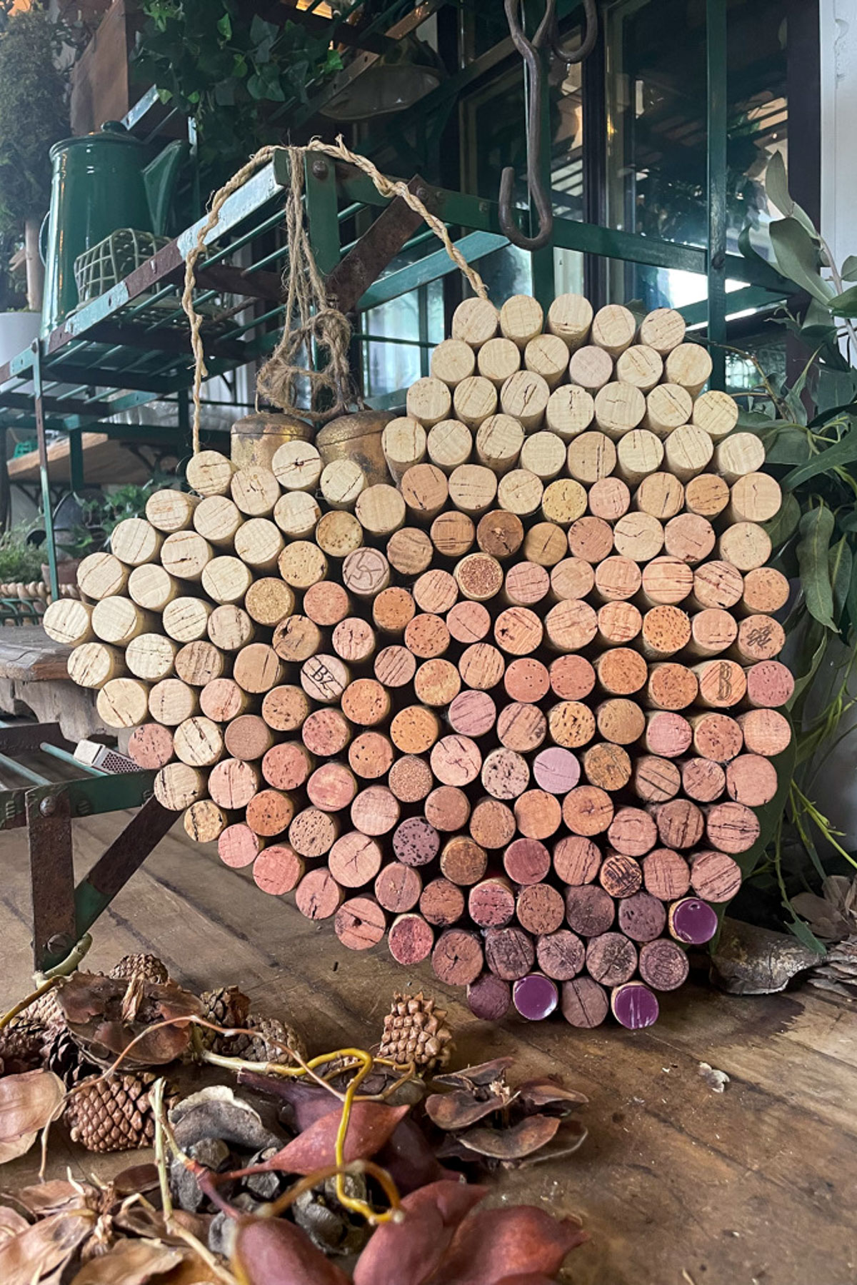 A heart-shaped decoration made of sliced branches in shades of brown and pink dangles from a twine loop. It's displayed on a wooden shelf, next to a rustic DIY wine bottle candle holder, surrounded by plants and natural materials like seed pods and dried leaves.