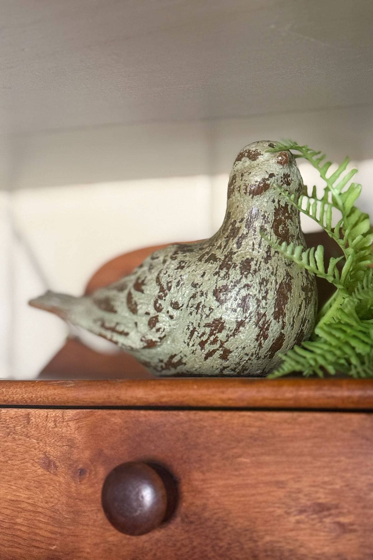 A small green and brown ceramic bird figurine sits on a wooden surface next to a fern leaf. The bird has a rustic, weathered appearance and the wooden surface features a round drawer knob.