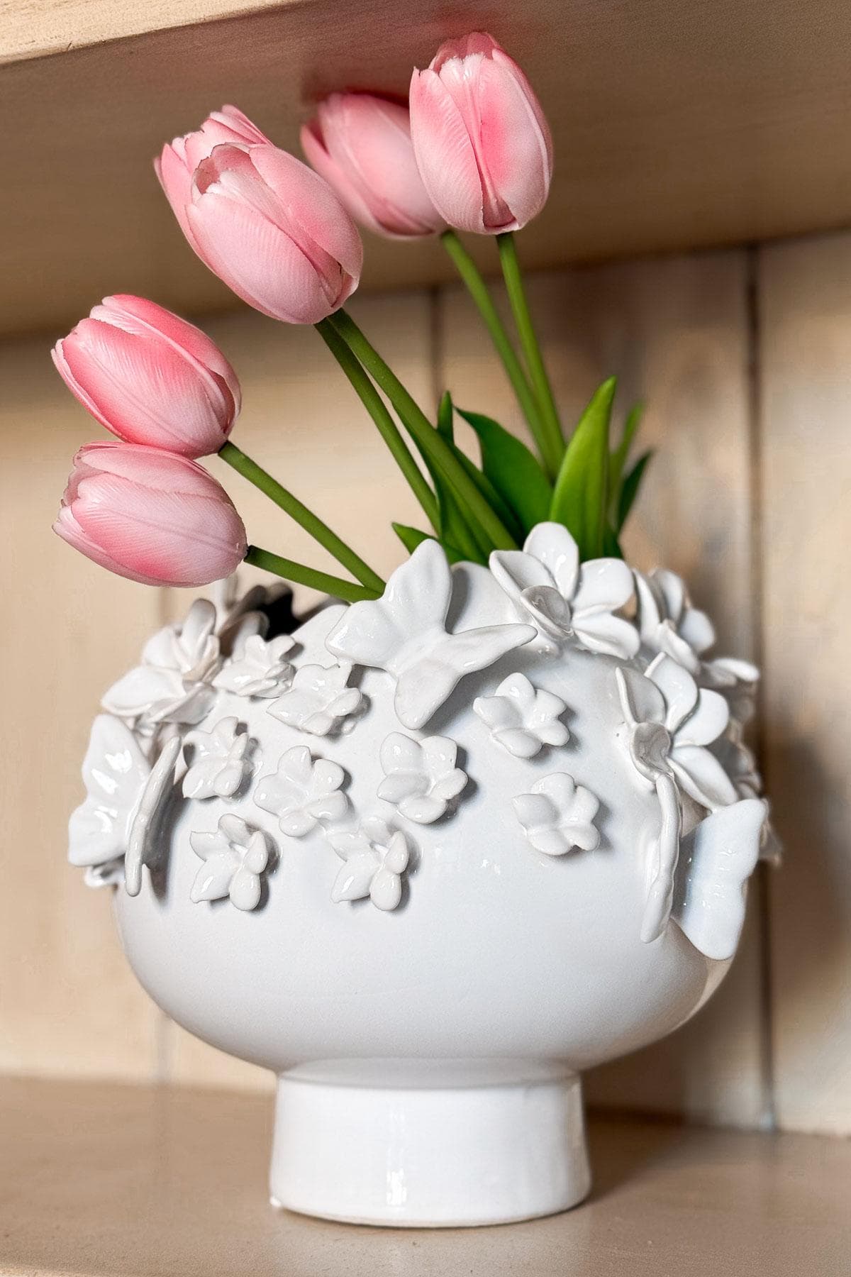 White ceramic vase adorned with floral designs, holding five pink tulips with green stems. The vase is placed on a wooden shelf, adding a decorative touch to the setting.