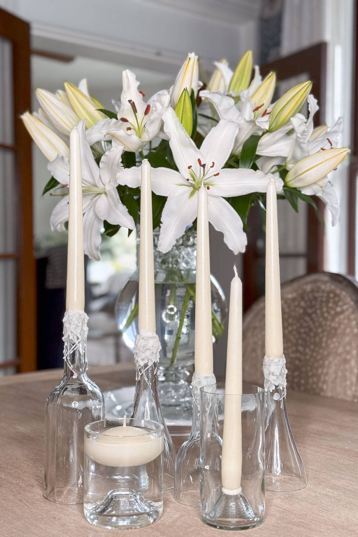 A dining table with a vase of white lilies in the background. In the foreground, glass bottles hold white taper candles of varying heights, and a small glass holds a white tealight candle. The scene is well-lit and elegant.