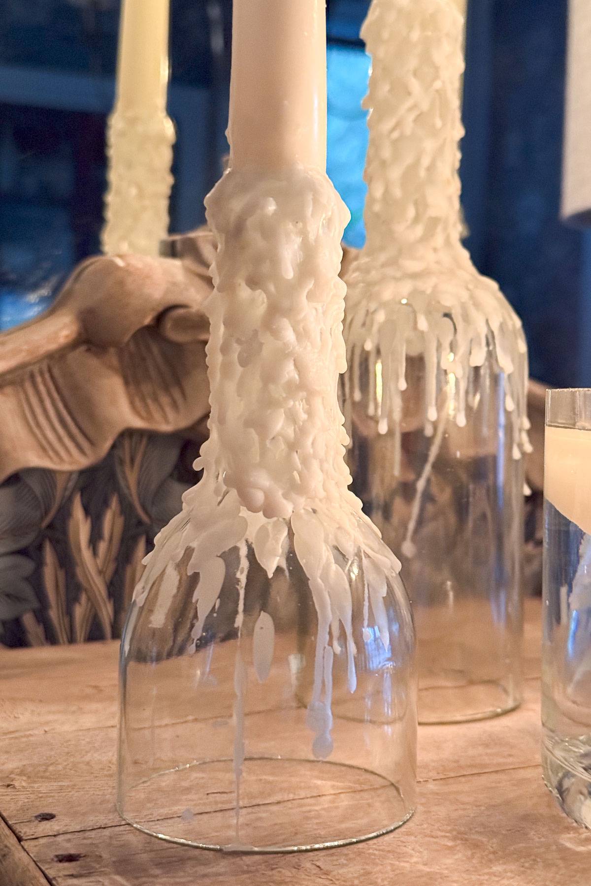 Glass bottles used as candle holders with white wax dripped down their sides, creating a textured, artistic effect. The bottles are placed on a wooden surface, and the scene is softly lit, highlighting the intricate wax patterns.