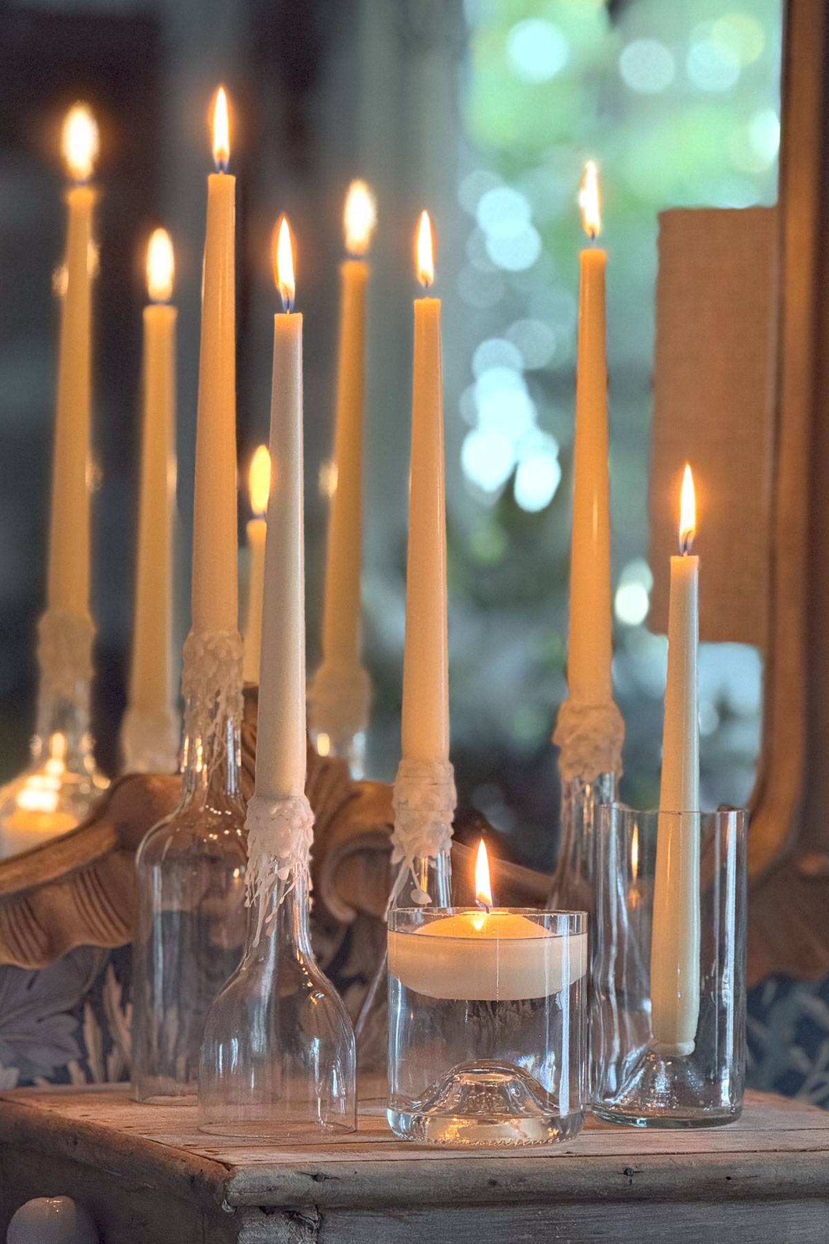 Tall white candles are lit and placed in clear glass bottles and holders on a wooden table. The setup is in front of a mirror, doubling the candlelight, with a blurred leafy background visible.