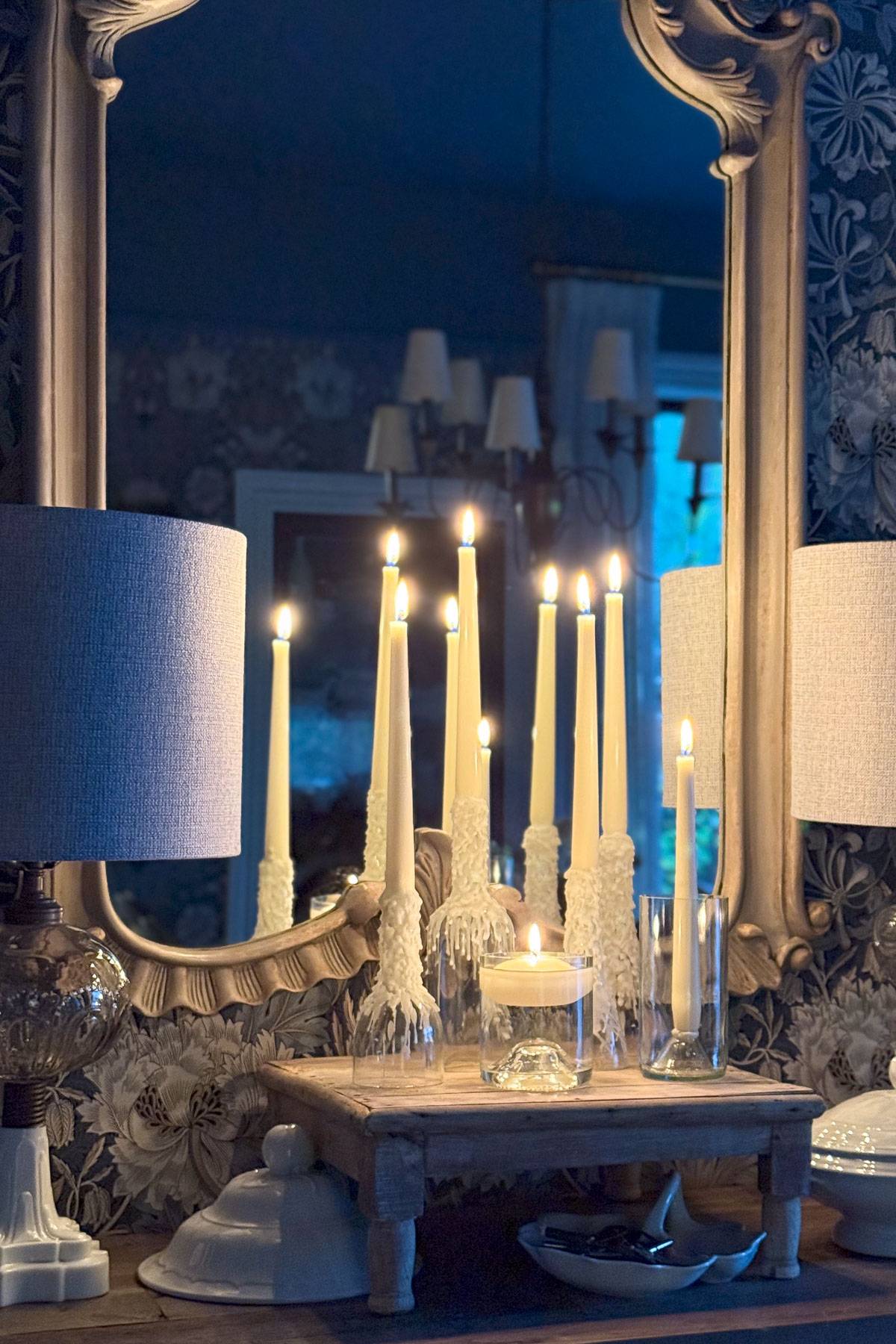 A decorative setting featuring tall, lit white candles on wooden stands in front of an ornate mirror. The reflection shows additional candles and a chandelier. The background includes a floral-patterned wallpaper and a pair of table lamps.