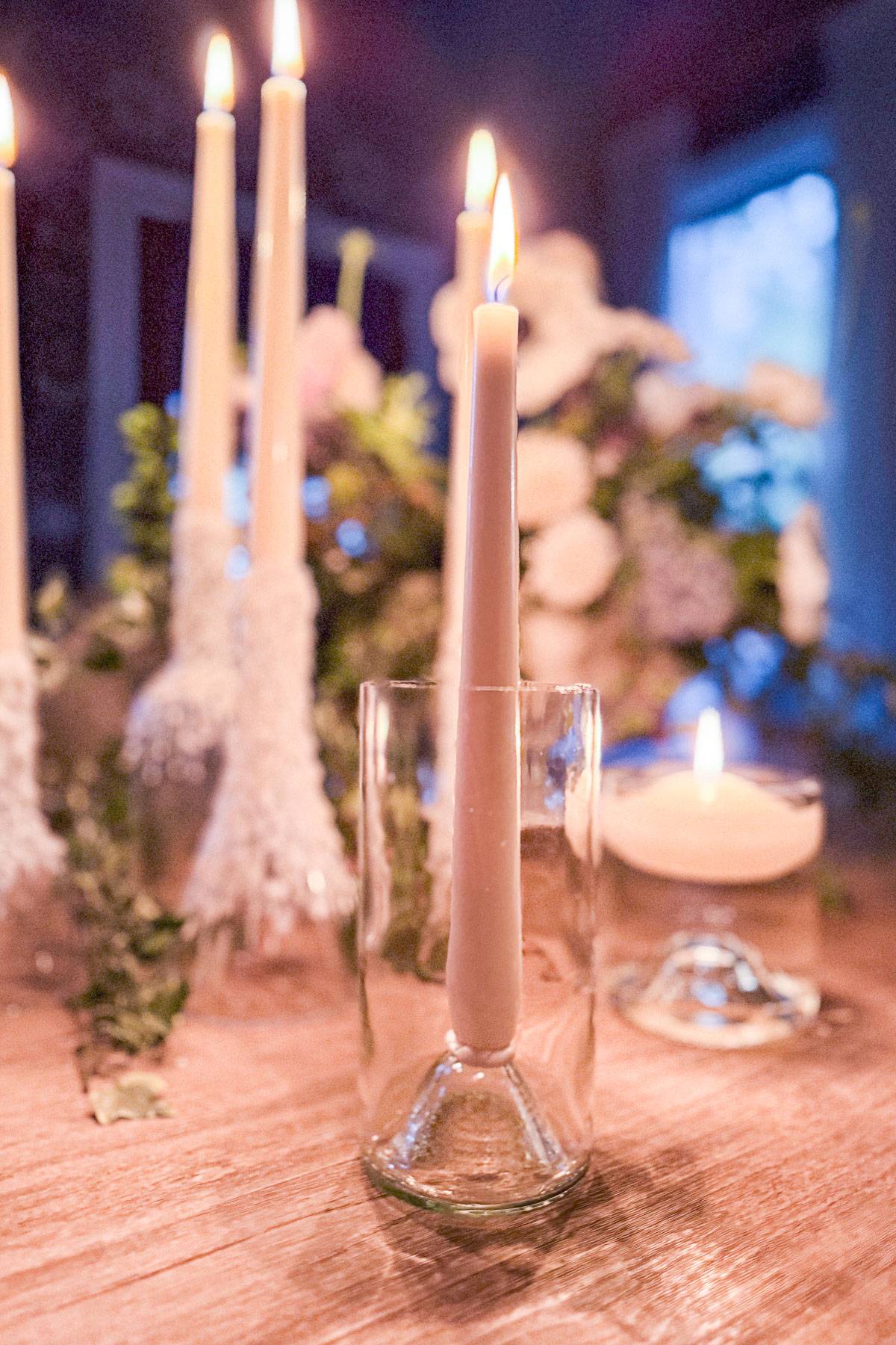 A lit candle placed inside a clear glass sits on a wooden table amidst other burning candles. In the background, blurred flowers and greenery contribute to a warm, ambient setting.