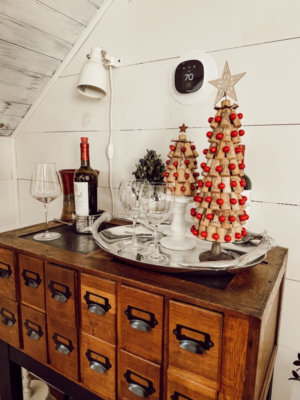 A rustic sideboard with labeled drawers showcases a tray featuring two decorative Christmas trees adorned with red beads, a DIY wine bottle candle holder, and four wine glasses. Above, a wall lamp and thermostat are mounted on the shiplap wall.