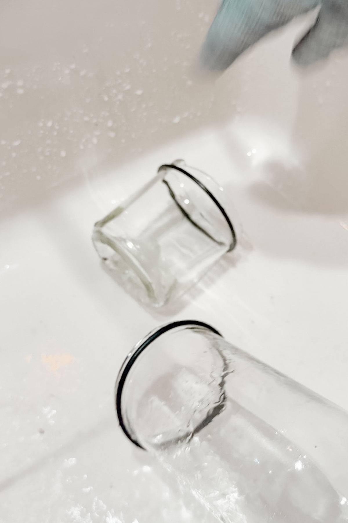 Two transparent glasses with black rims are lying horizontally in a white sink, partially submerged in water. A gloved hand is visible in the top right corner, suggesting the action of washing the glasses.