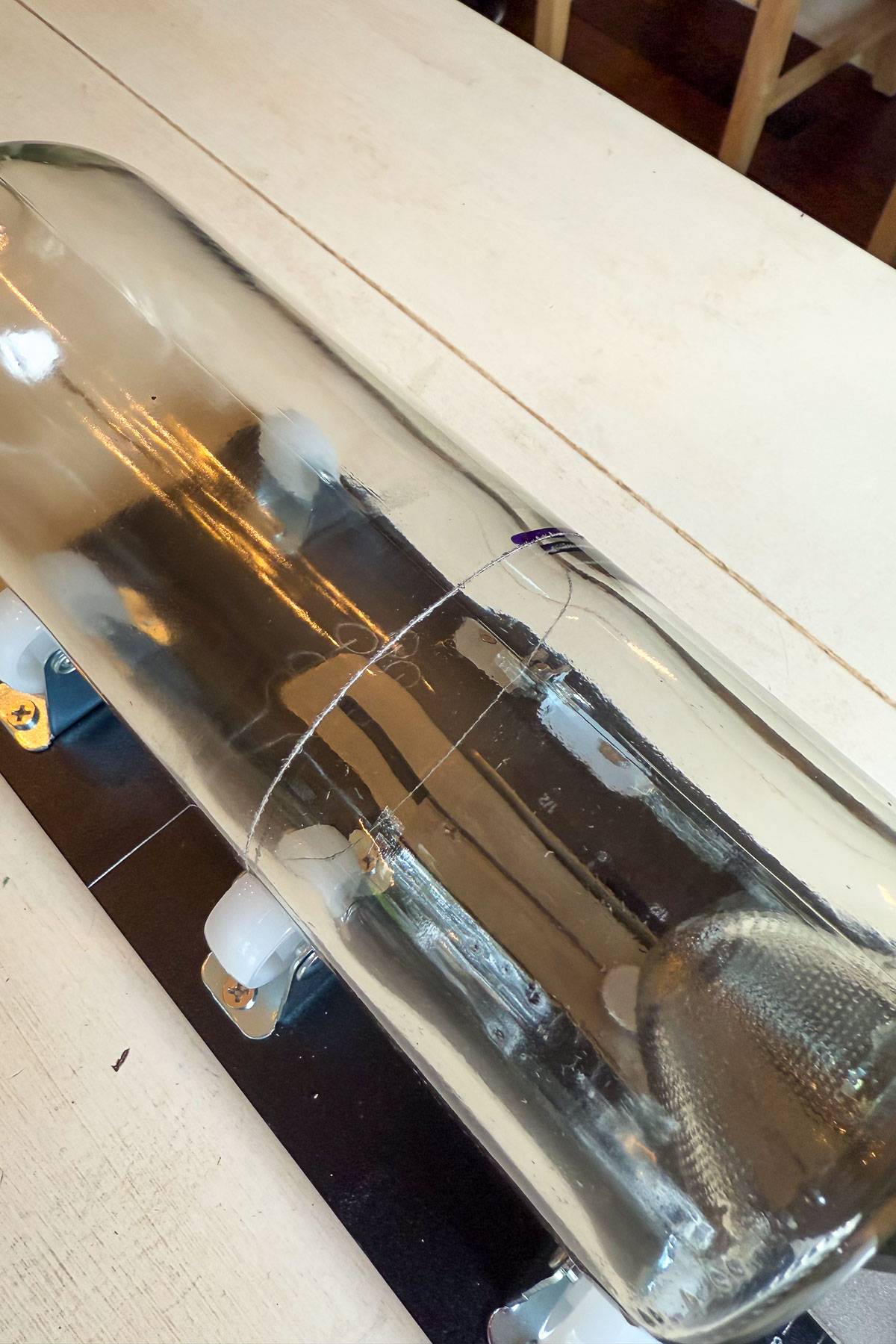 A large, clear glass bottle is placed horizontally on a white table with a string wrapped around its middle, possibly as a cutting guide. The bottle is supported by a makeshift holder with white wheels.