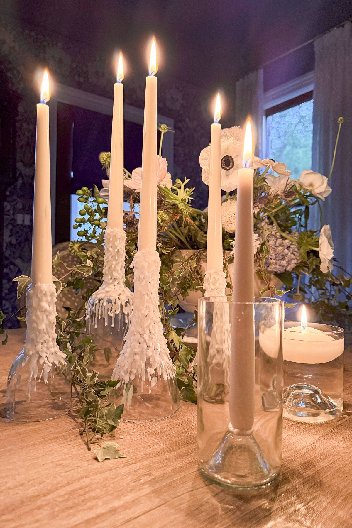 Candles in glass bottles, with wax dripped decoratively over them, are lit on a wooden table. A small, lit candle in a clear glass holder sits nearby. In the background, a floral arrangement with white and green flowers is visible.