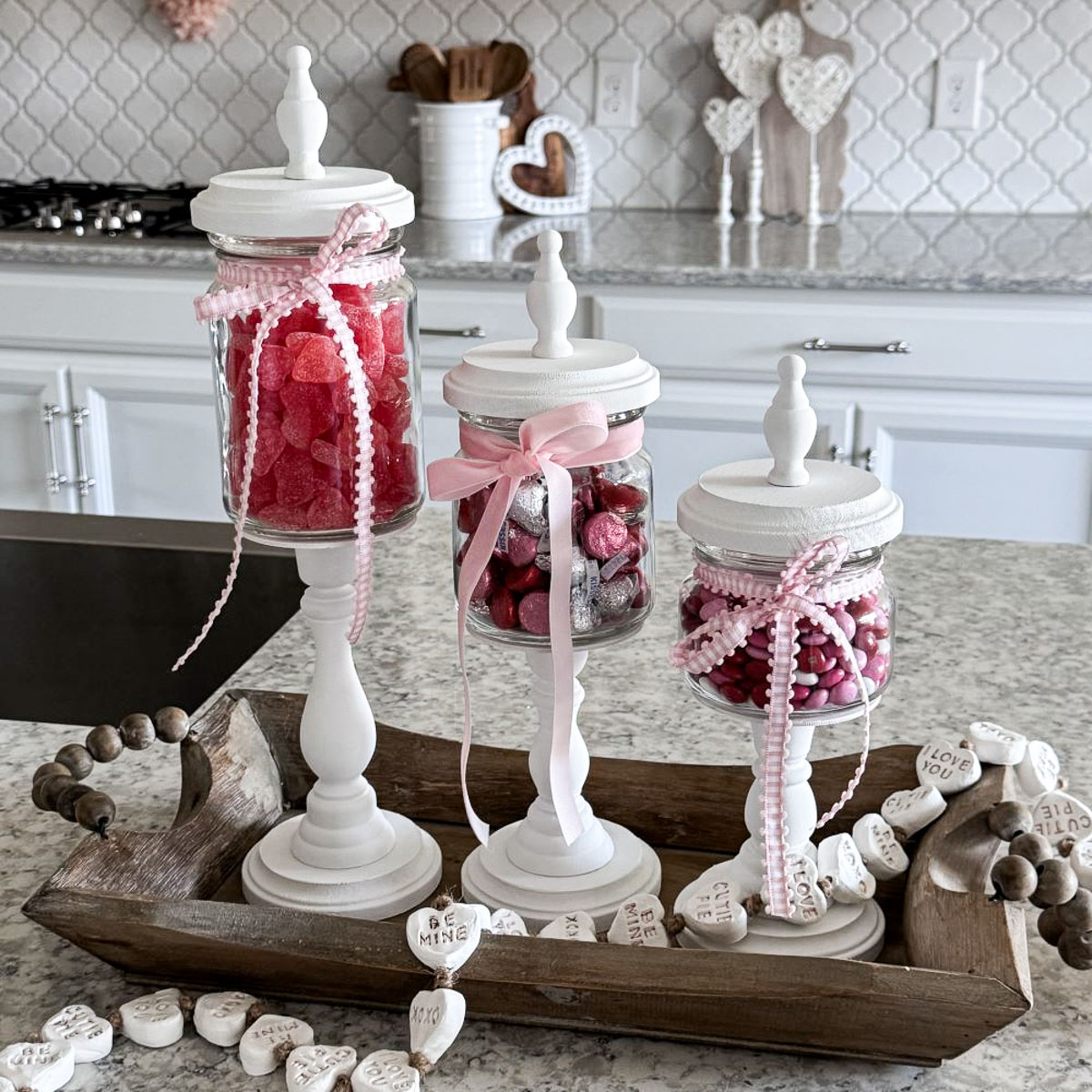 Three jars filled with red and pink candies, adorned with ribbons, are displayed on a kitchen counter. They rest on a wooden tray, surrounded by heart-shaped decorations, in a kitchen setting with a white backsplash and countertops.