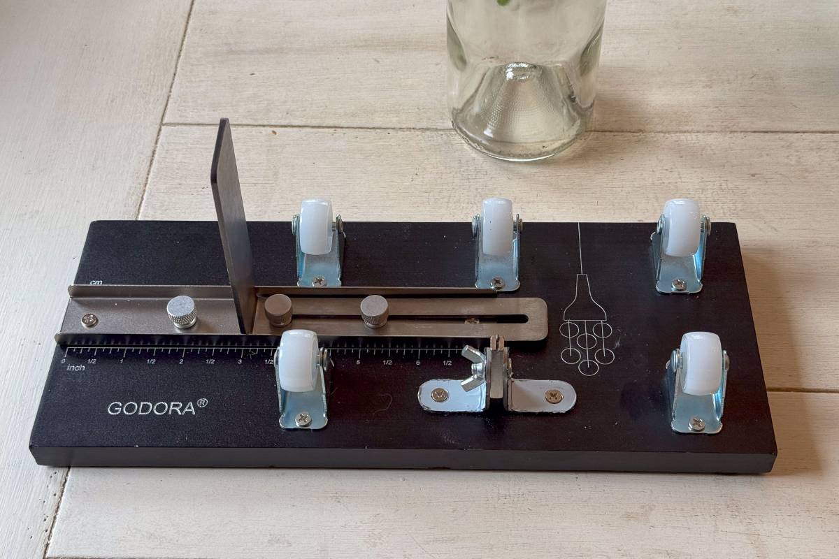 A glass bottle cutter sits on a wooden table. The device features a black base with adjustable metal components and five white wheels. A clear glass bottle is partially visible at the top of the image.