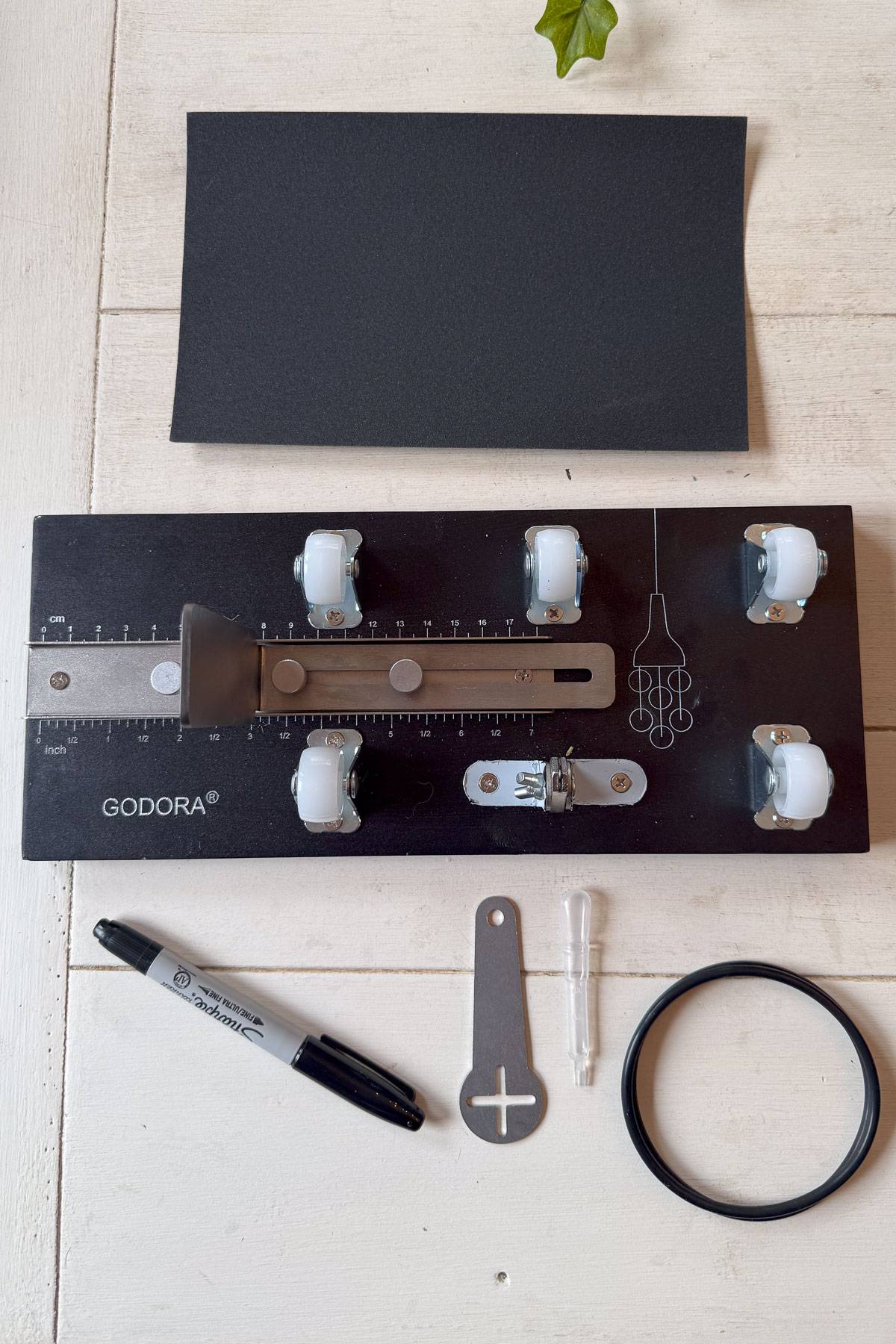 A crafting kit with various tools arranged on a wooden surface. Includes a black board with rollers, a marker, a metal tool, a small transparent container, a rubber band, and a black sheet. A small green leaf is partially visible at the top.