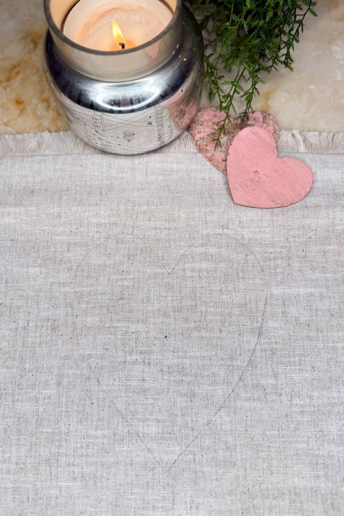 A lit candle in a silver holder sits on a cream-colored surface next to a sprig of greenery. Two heart-shaped cutouts, one pink and one faintly outlined on fabric, are placed beside the candle.