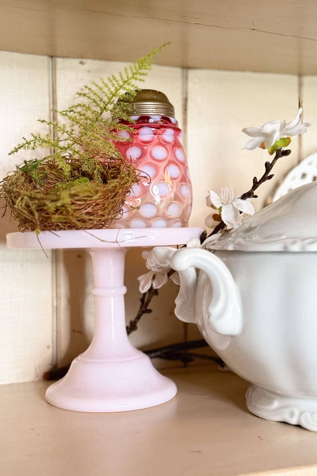 A decorative display featuring a small bird's nest on a pink pedestal stand with a red and pink textured glass jar. Beside it, there's a white ornate teapot with a branch of white flowers resting on it.