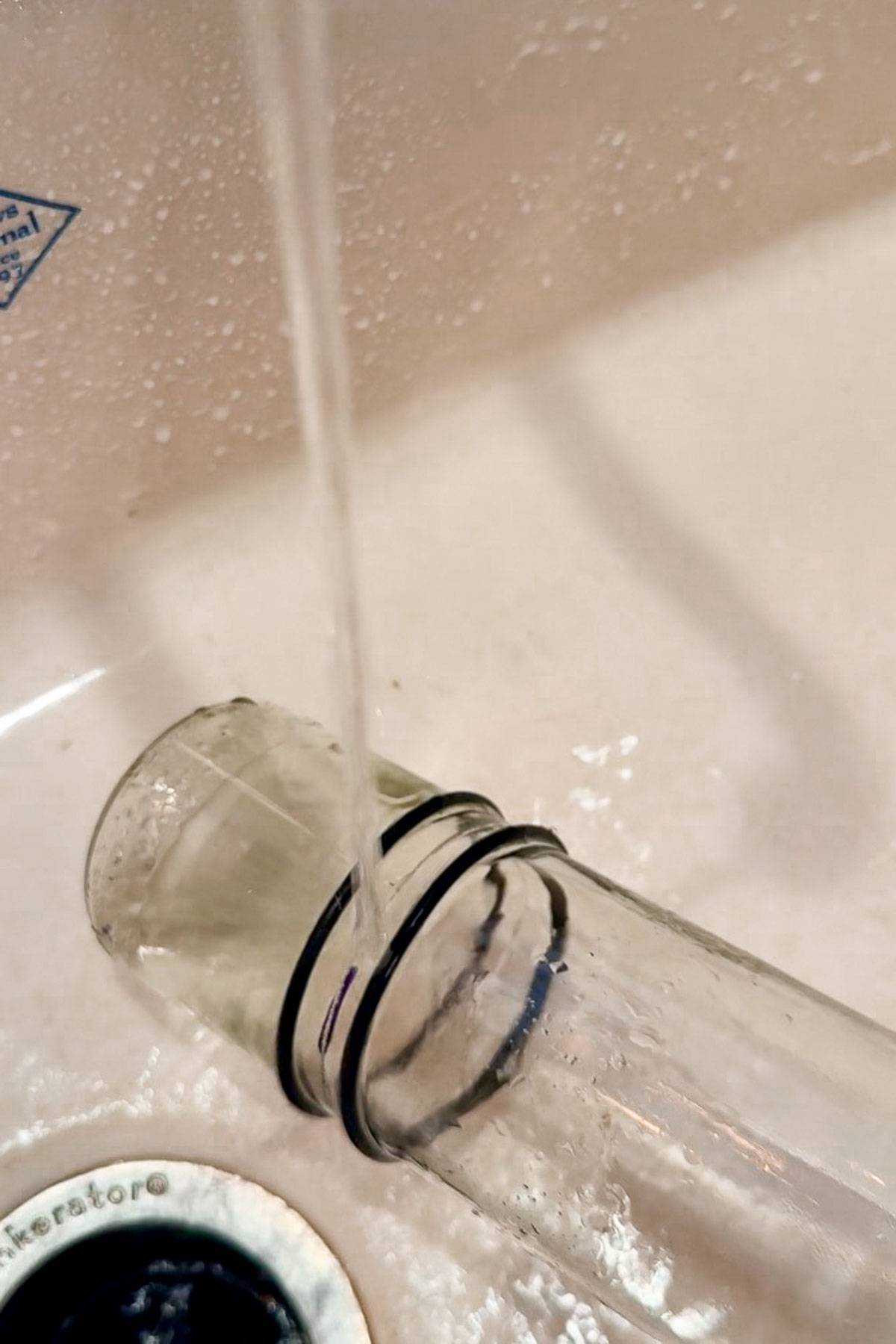 A clear glass container with black rubber rings is being rinsed under a stream of water in a white sink. The sink's metal drain is visible in the bottom left corner. Water droplets are scattered around the sink's surface.