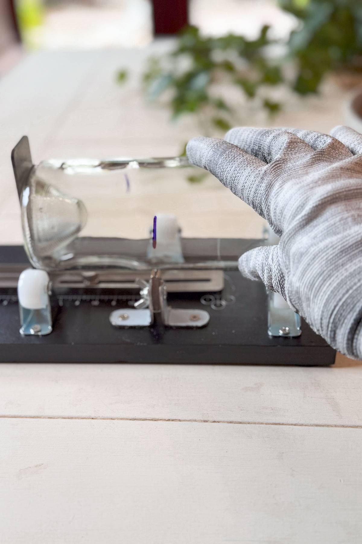 A gloved hand stretching a transparent, flexible sheet over a metal clamp set on a wooden surface. A plant is blurred in the background. The scene suggests a science experiment or demonstration.