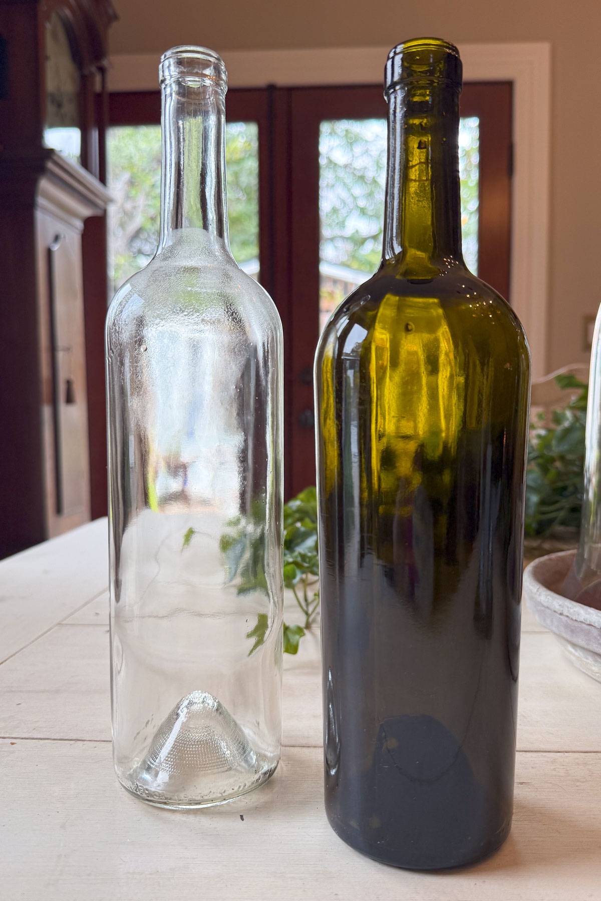 Two empty wine bottles on a table: one clear and one green. The background features a pair of glass doors with greenery visible outside.