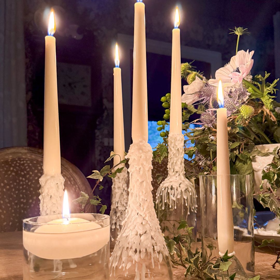 Lit candles of varying heights, with wax drips cascading down the sides, are arranged on a table. A glass vase with delicate white and purple flowers sits nearby. The scene has a warm, cozy ambiance.