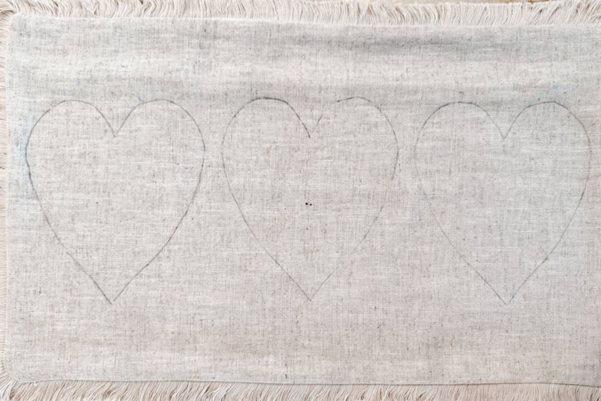 A textured beige fabric with three faintly outlined heart shapes drawn side by side. The fabric features fringed edges at the top and bottom.