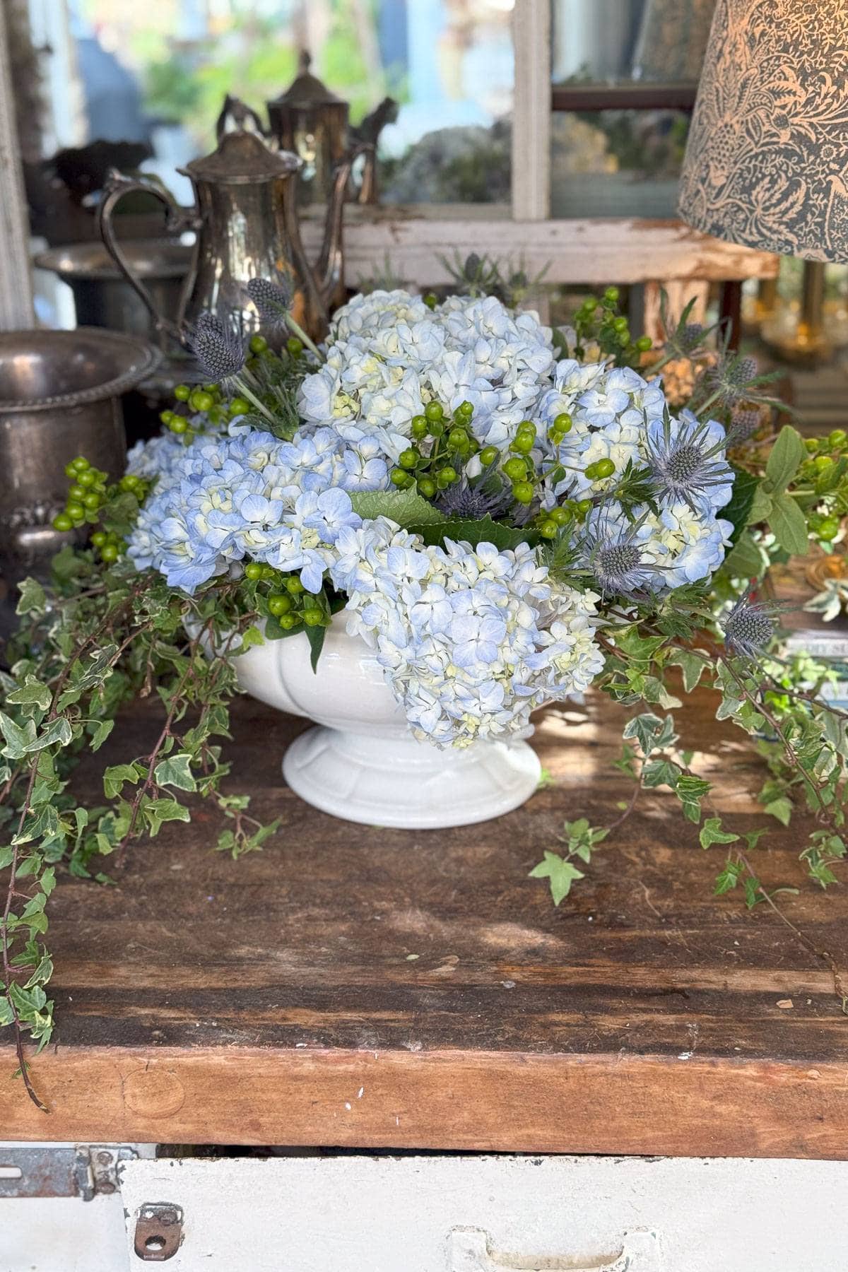 A rustic floral arrangement featuring light blue hydrangeas, green berries, and ivy in a white ceramic vase on a wooden table. Behind the bouquet are antique silver pitchers and a patterned lampshade, creating a vintage ambiance.