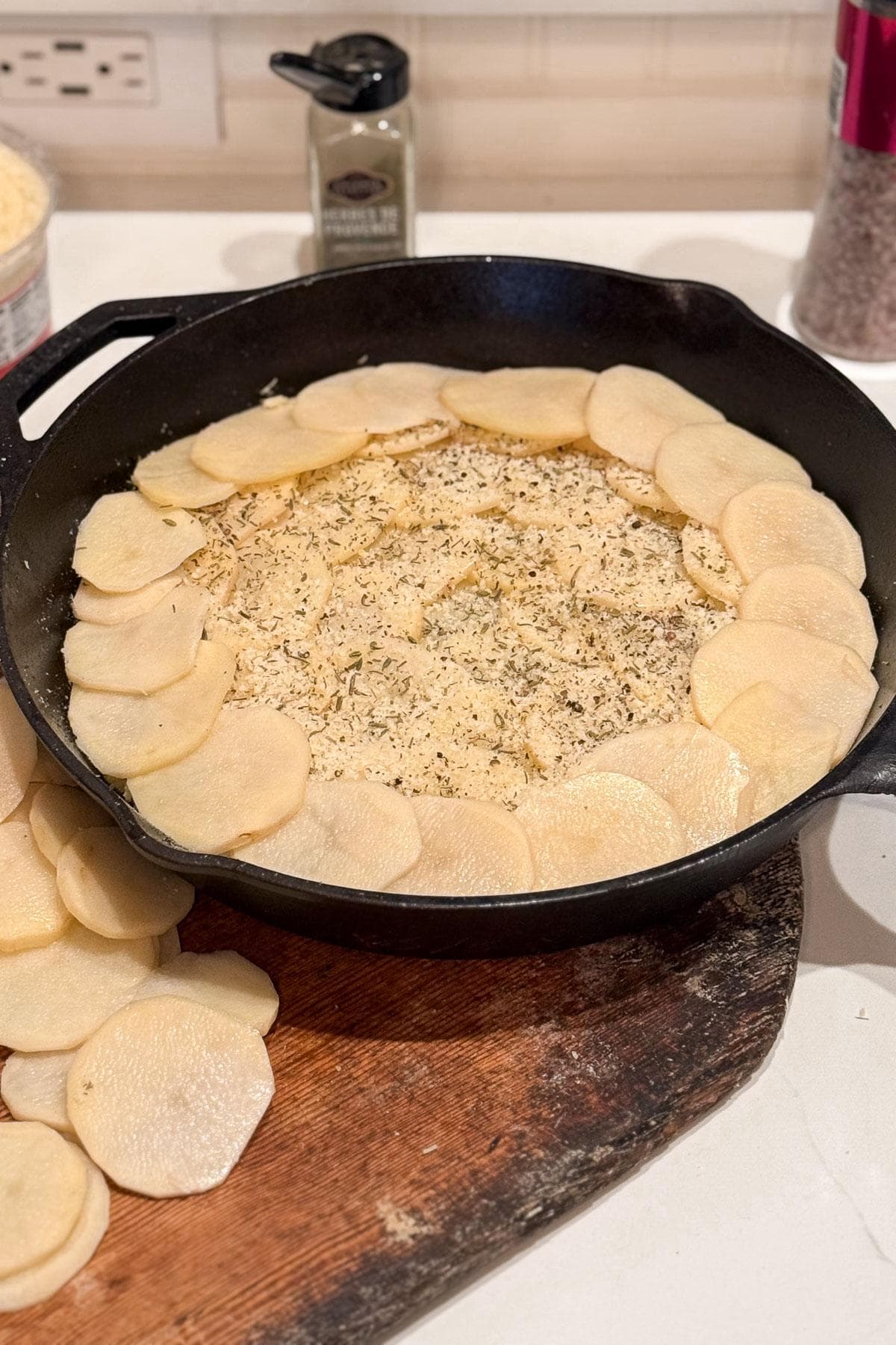 Making layered potato bake. Sliced potatoes are arranged in a circular pattern around the edge of a cast iron skillet, with grated cheese and herbs in the center. Extra potato slices are on a wooden cutting board nearby. Cheese container is in the background.