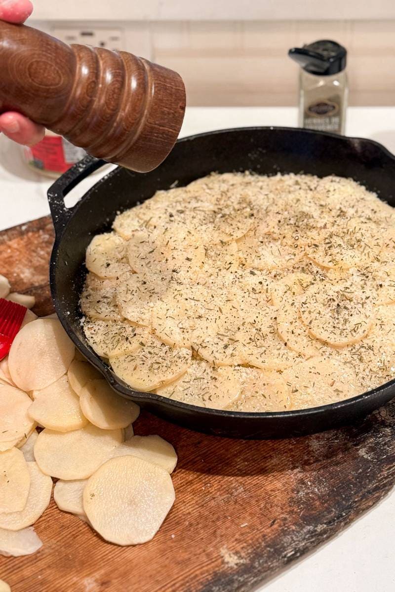 A hand holding a wooden pepper grinder sprinkles pepper over sliced potatoes in a cast iron skillet to make layered potato bake. The skillet is on a wooden board, with extra potato slices beside it. A spice container is in the background.