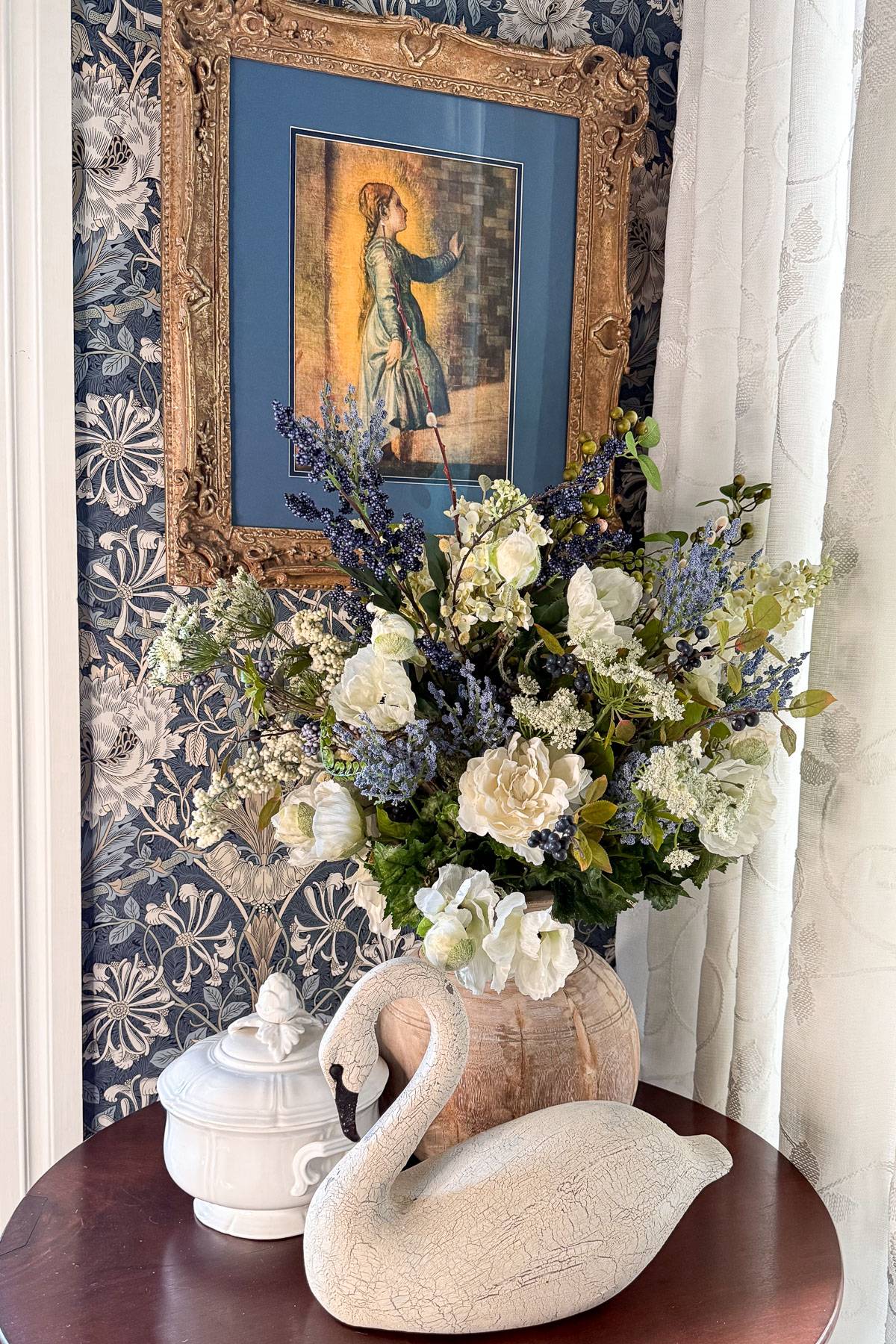 A table with a white swan-shaped ornament, a vase of white and purple flowers, and a white ceramic dish with a lid. Behind is a framed painting of a woman, set against floral-patterned wallpaper.