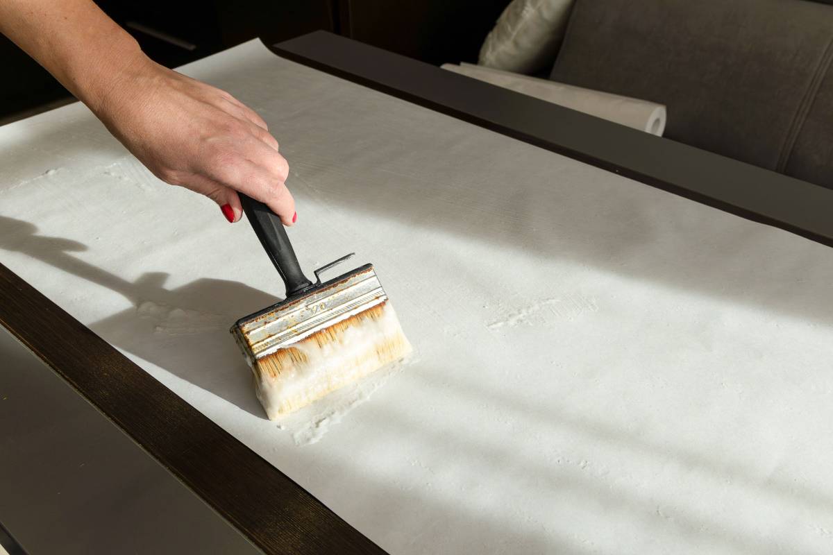 A hand uses a large brush on a flat, white surface, possibly preparing or cleaning the area. Light and shadow create patterns across the surface. A roll of paper is visible in the background.