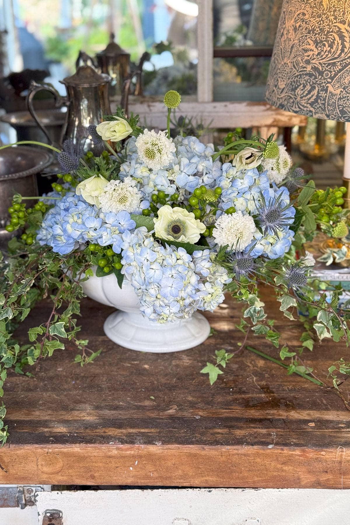 A white ceramic vase holds a floral arrangement with light blue hydrangeas, white roses, and greenery. It sits on a rustic wooden table, surrounded by ivy. In the background are silver teapots and a patterned lamp.