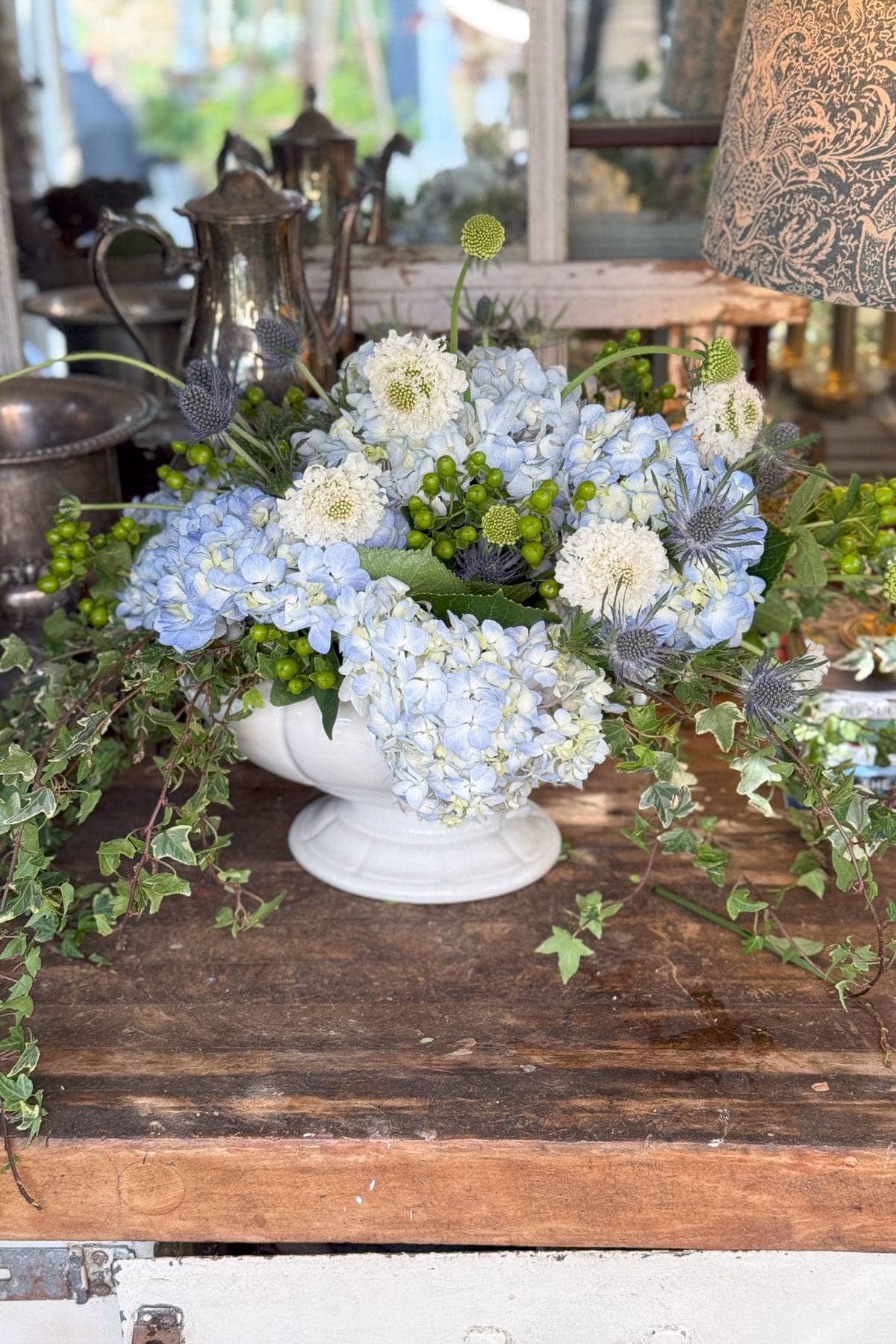 A floral arrangement in a white vase, featuring blue hydrangeas, white scabiosa, and green berries, sits on a wooden table. Ivy drapes over the edges. Behind are vintage silverware, a patterned lamp, and a glass-paned cabinet.