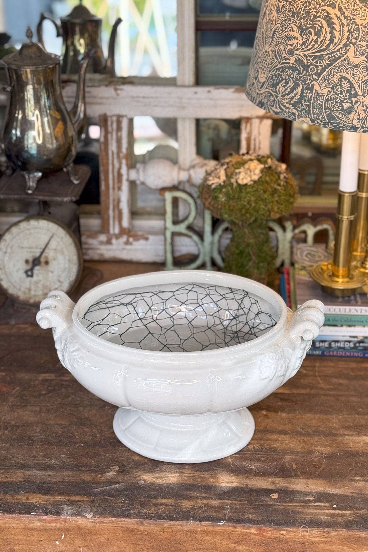 White ceramic bowl with two handles, featuring black wire mesh inside. It sits on a wooden table alongside vintage decor, including a silver teapot, an old scale, a floral-patterned lamp, and decorative books.