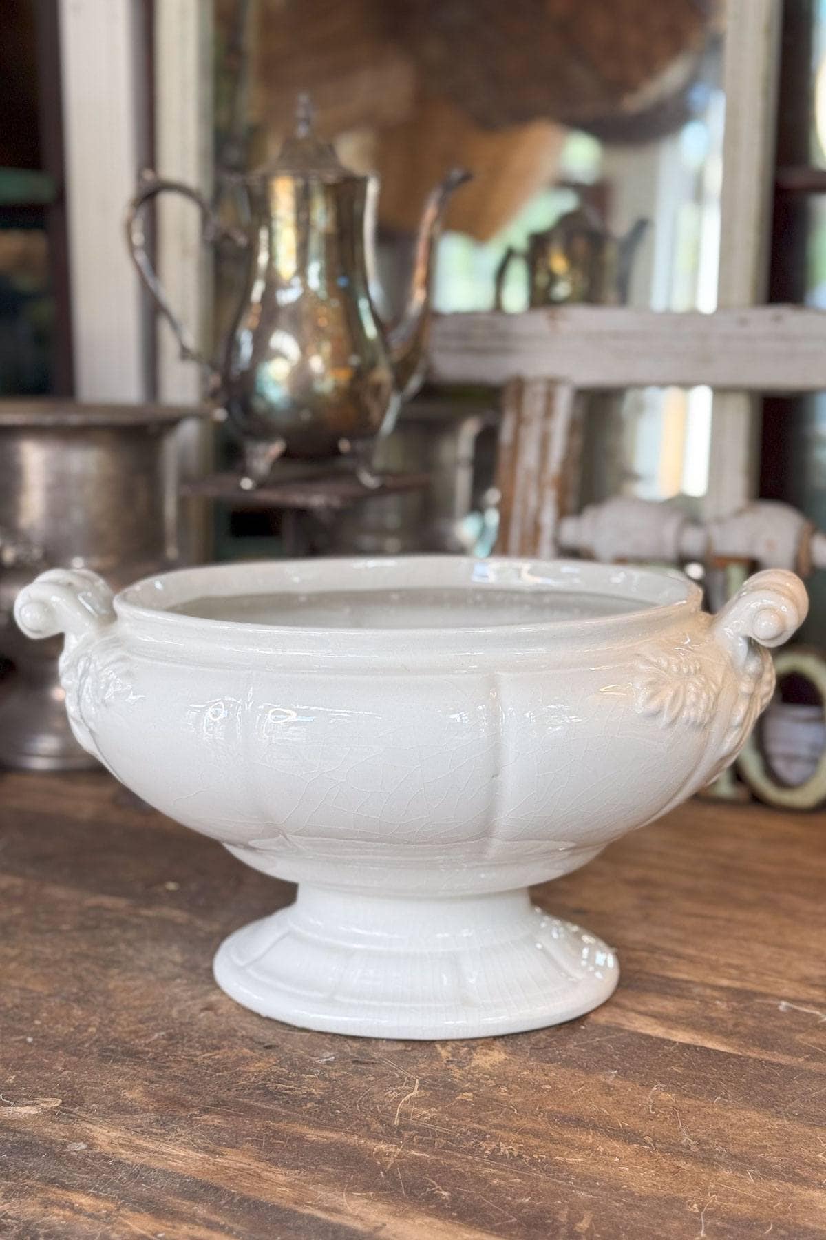 A white ceramic tureen with ornate side handles sits on a wooden surface. A silver teapot is blurred in the background, along with other vintage kitchen items, creating an antique kitchen atmosphere.