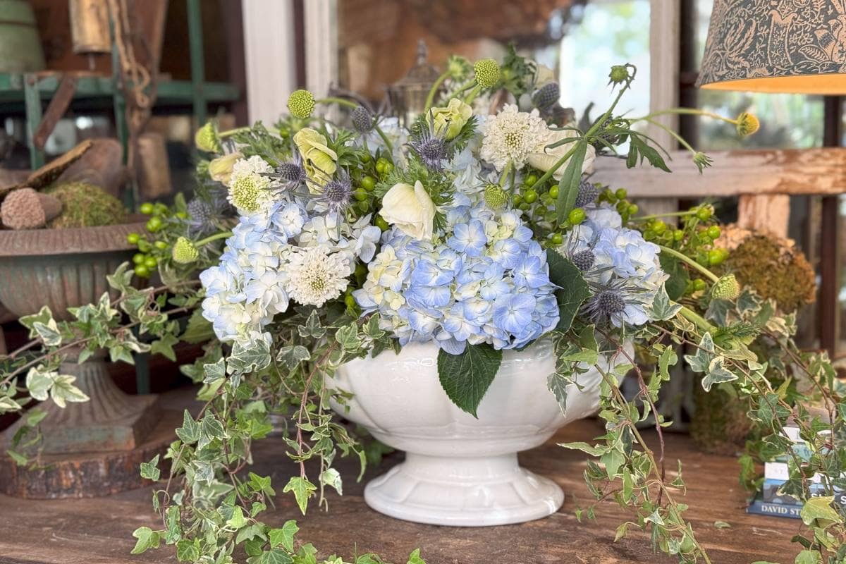 A floral arrangement sits on a wooden table, featuring blue and white hydrangeas, white roses, greenery, thistles, and cascading ivy in a white bowl. The background includes rustic decor elements and a lampshade.