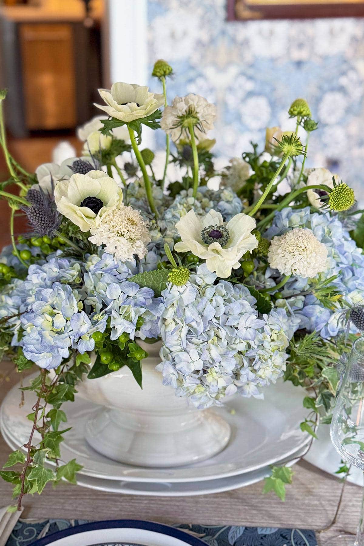 A floral arrangement featuring white and light blue flowers, including hydrangeas and anemones, in a white vase. The vase sits on a white plate with ivy draped around the base. The background is softly blurred, with floral-patterned wallpaper visible.