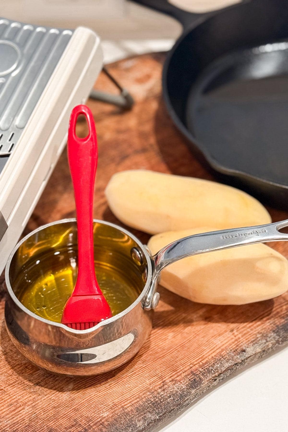 A small saucepan filled with oil and a red silicone brush rests on a wooden board. Two peeled potatoes are nearby, with a waffle maker and a cast iron skillet in the background.