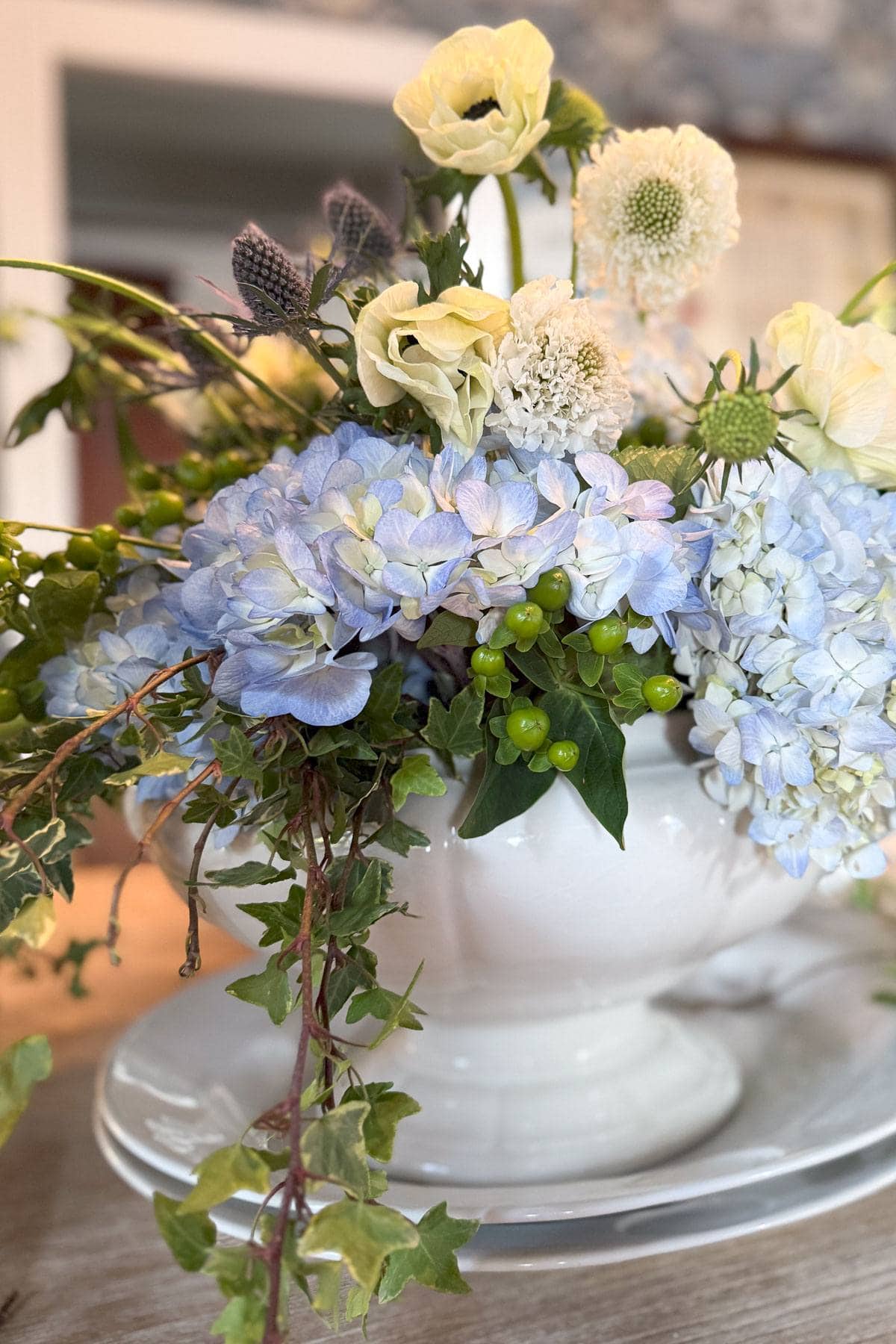 A floral arrangement in a white vase features light blue hydrangeas, white blooms, green berries, and ivy, set on a white plate. The background is softly blurred, highlighting the elegant and fresh arrangement.