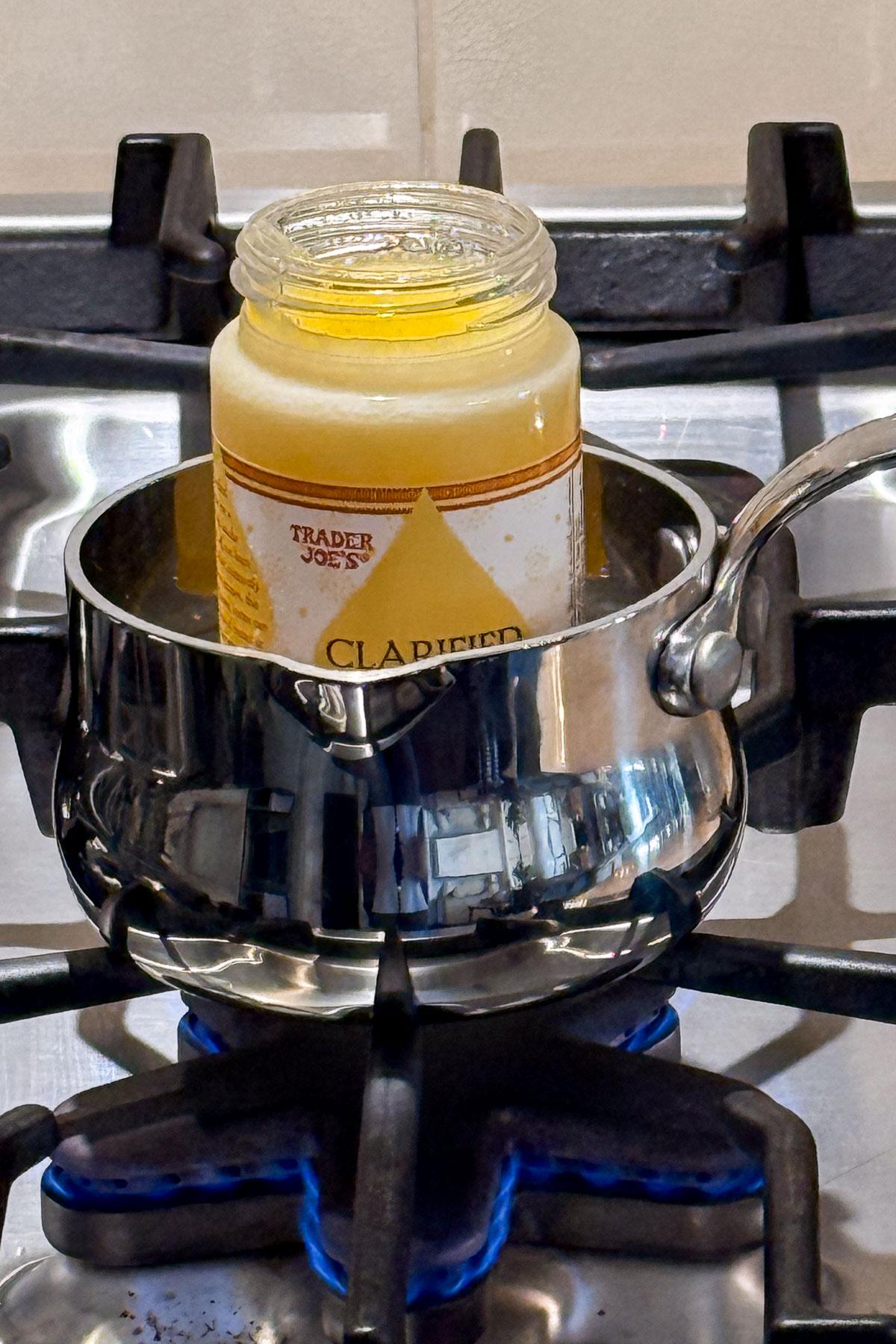 A jar of Trader Joe's clarified butter is being heated in a small stainless steel pot on a gas stove. The blue flame is visible beneath the pot.