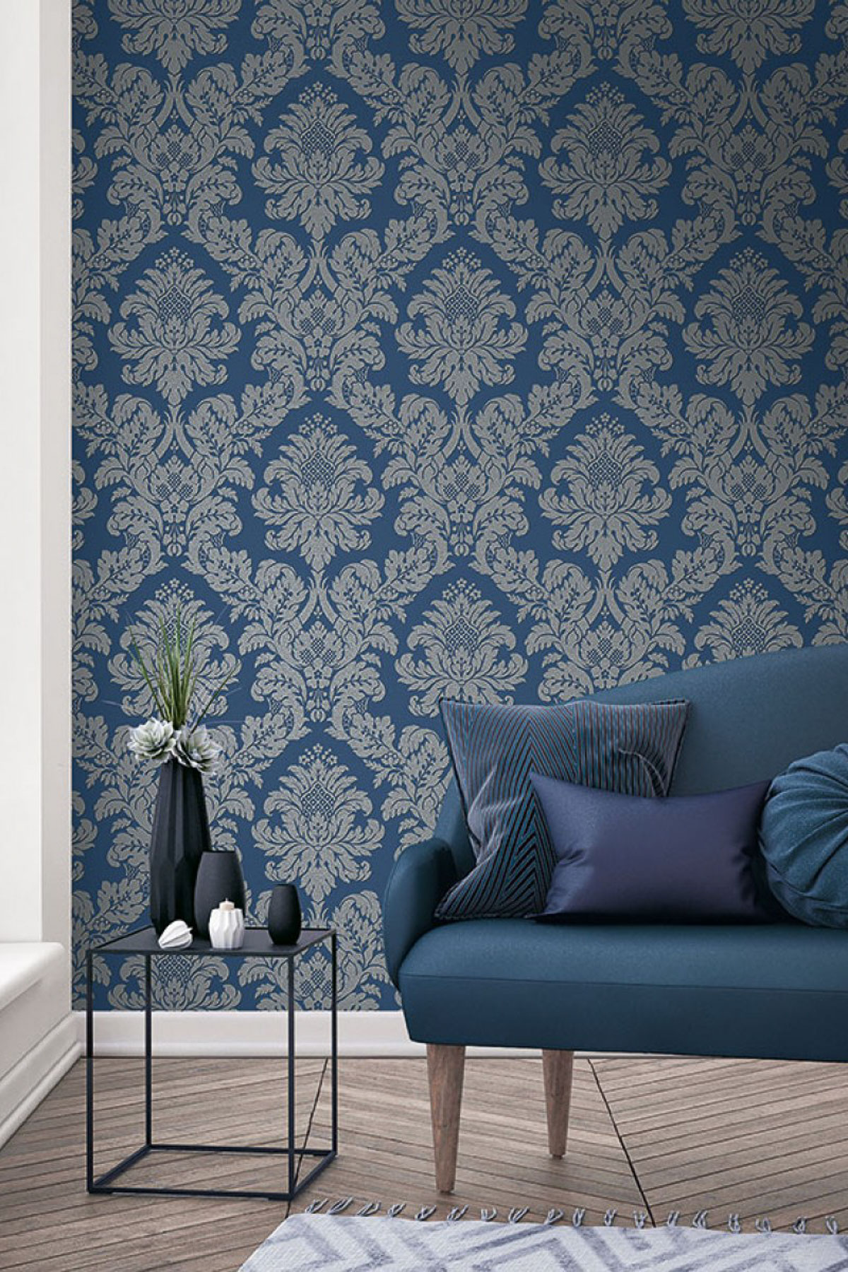 A room with a blue damask patterned wallpaper features a plush blue sofa adorned with pillows, while a side table holds vases with flowers. The light wood floor complements the space, and natural light streams through a window on the left, creating an inviting atmosphere.