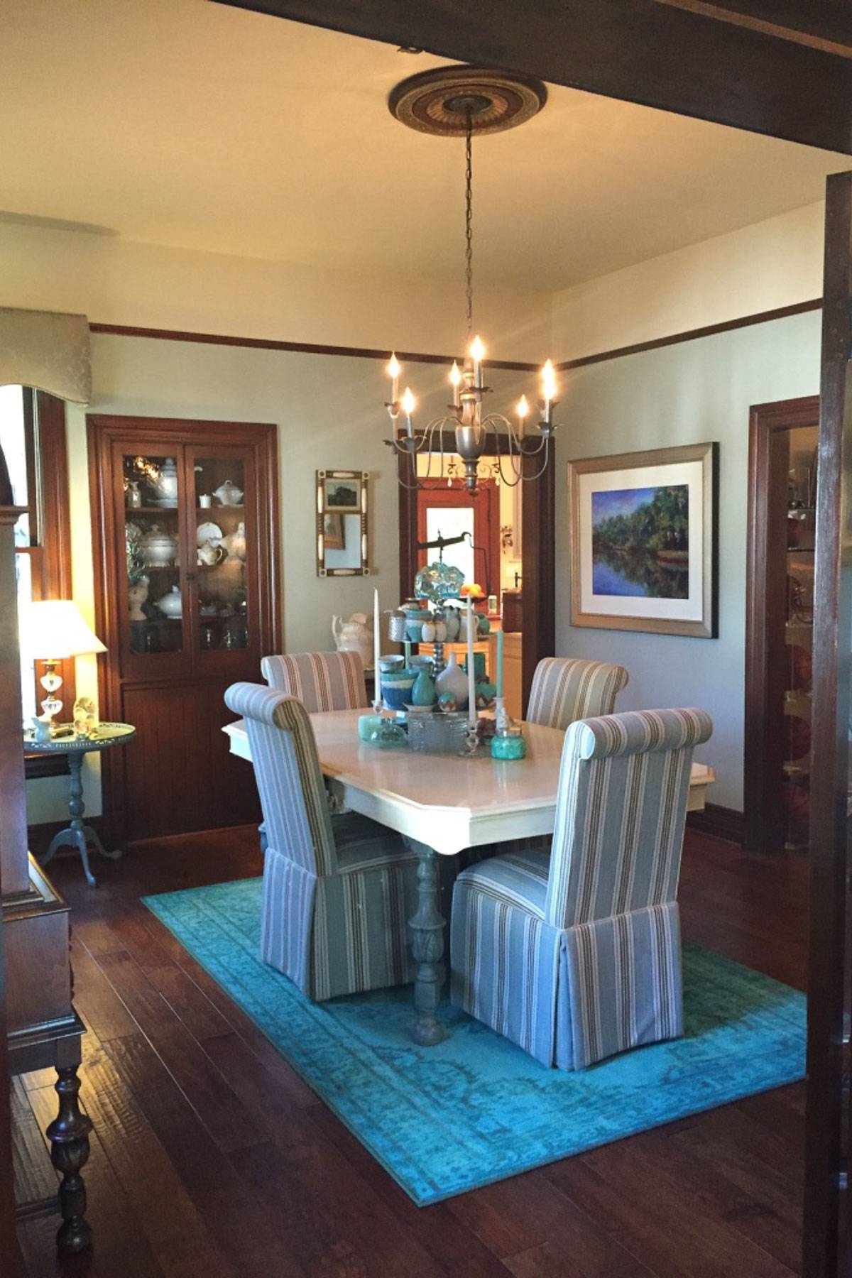 A cozy dining room with a white square table surrounded by striped cushioned chairs. A decorative chandelier hangs above. The room features wooden floors, a blue rug, a corner cabinet with dishes, and a landscape painting on the wall.