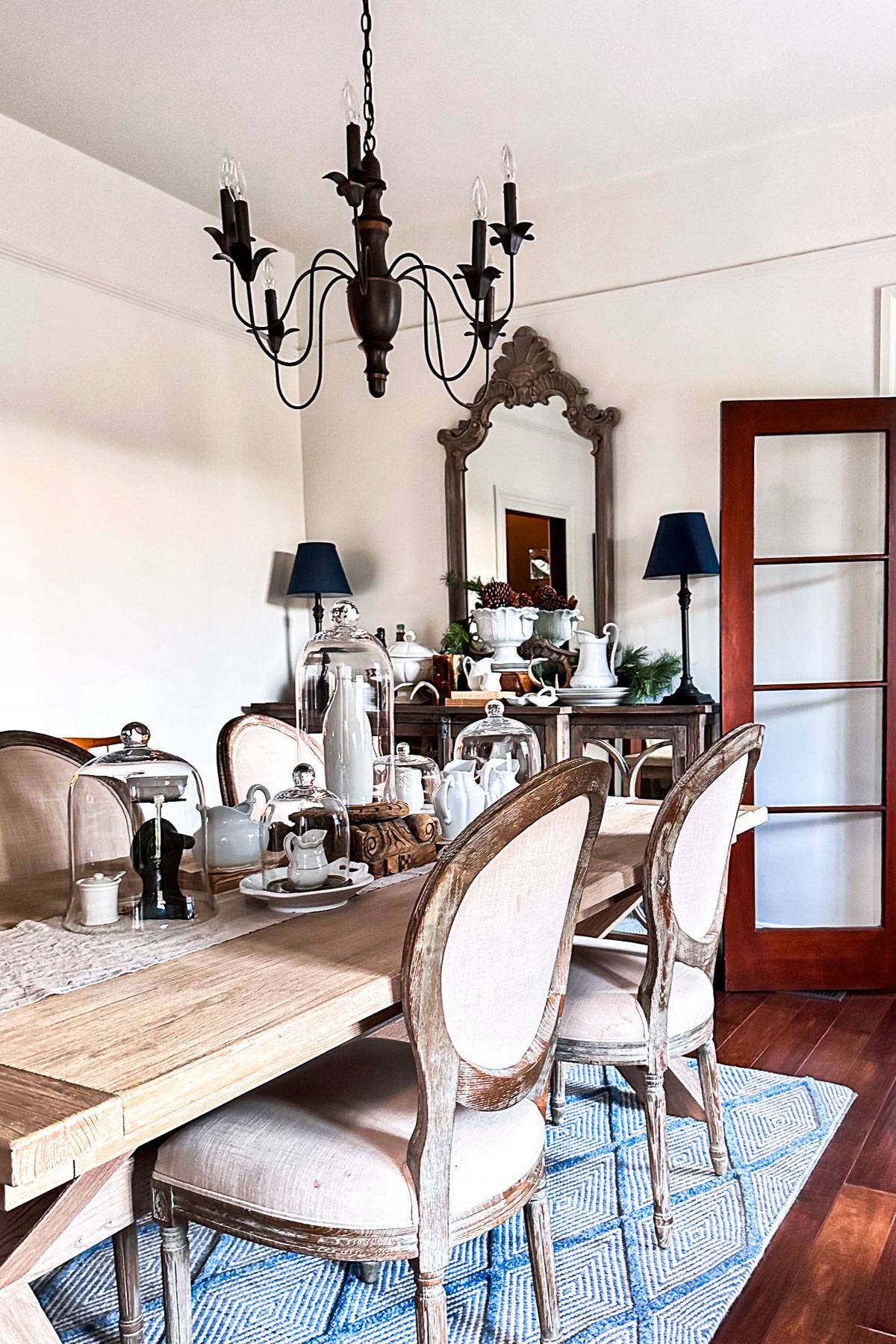 A cozy dining room with a wooden table and six rustic chairs on a patterned rug. A black chandelier hangs above. The back wall has a decorative mirror, a sideboard with lamps, and various decorative objects. A door with glass panels is visible.