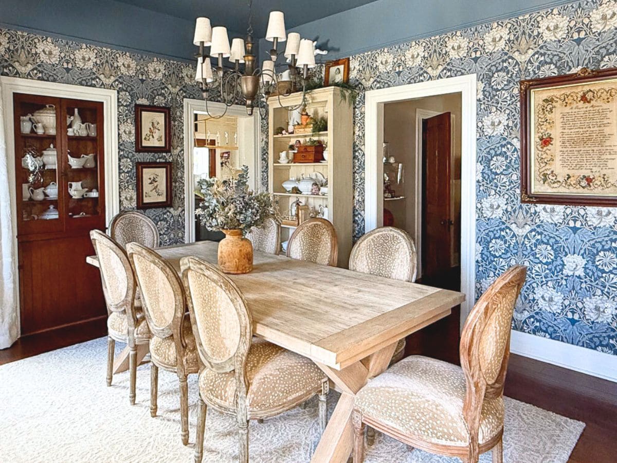 Updating a dining room with floral wallpaper, this space features a wooden table and eight cushioned chairs beneath a shimmering chandelier. A cabinet displays elegant dishware, while framed art adds charm to the blue patterned walls. A light carpet completes the inviting ambiance.
