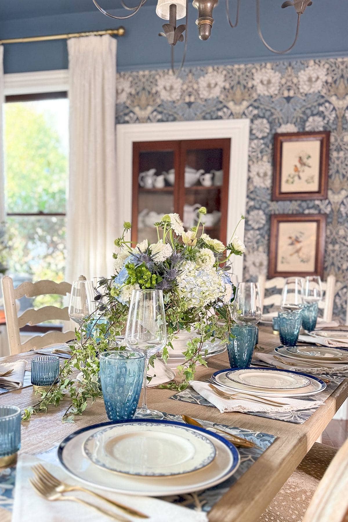 A dining table is elegantly set with blue and white plates, clear glassware, and blue water glasses. A floral centerpiece with white and blue flowers sits in the middle. The room has a floral wallpaper and a glass cabinet in the background.